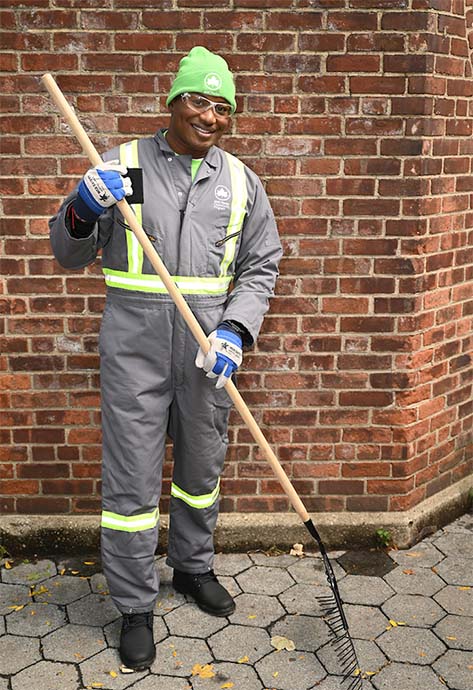 A worker wearing a clean grey parks uniform and a stylish lime green beanie holds a rake. He is outdoors and looks content.