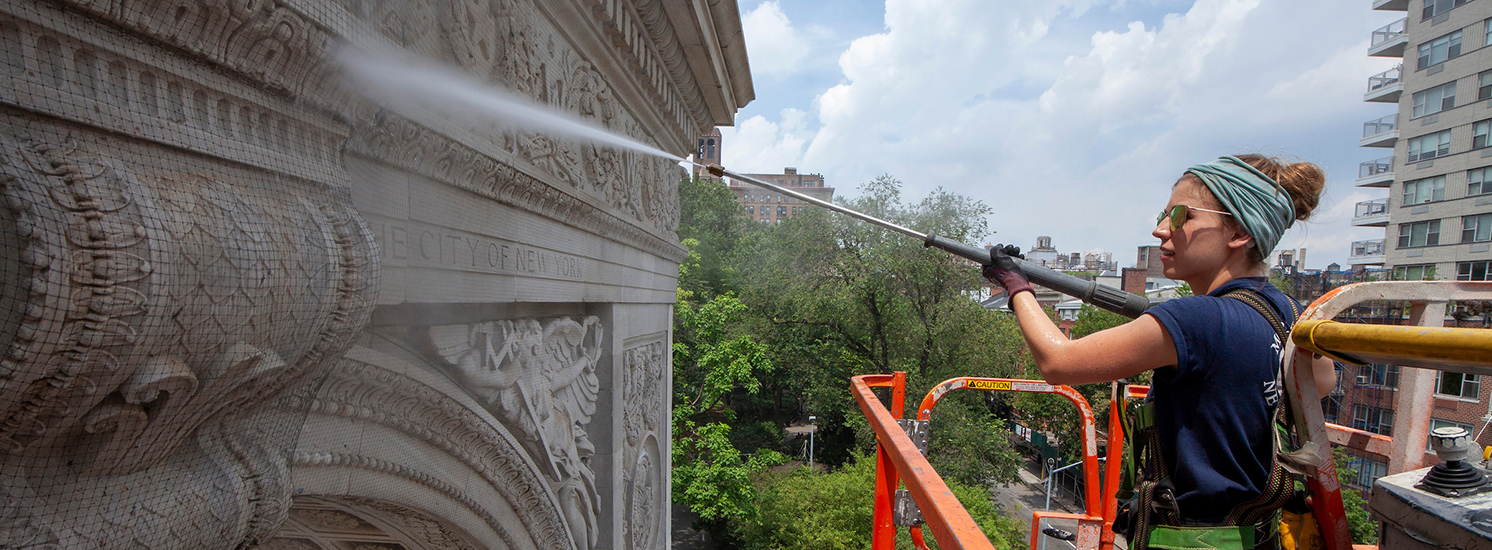 A woman in a forklift power washes the recognizable Washington Square Arch.