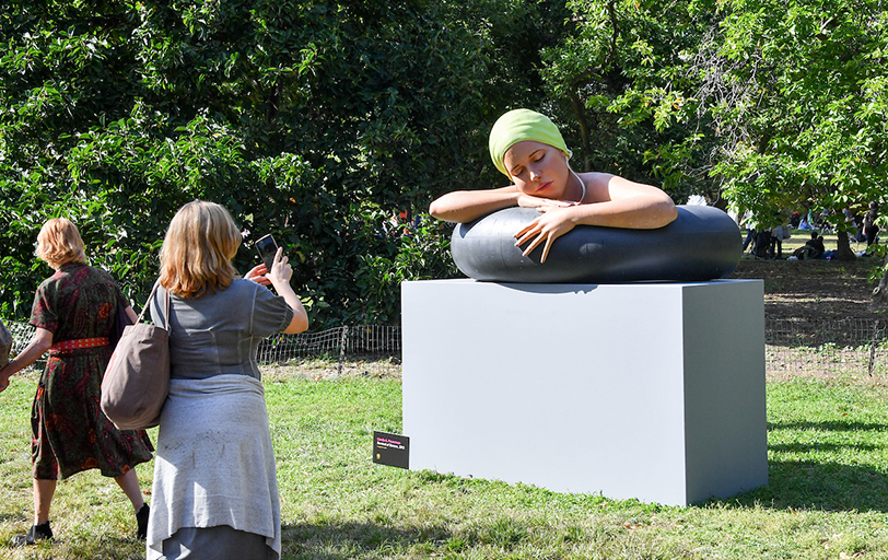 A woman takes a photo of a realistic bust that artistically depicts a woman sleeping on an inner tube in a pool.