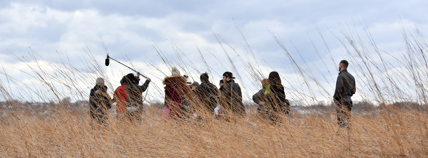 People wearing winter clothes stand in grasses. One person is holding a boom mic to indicate recording is taking place.