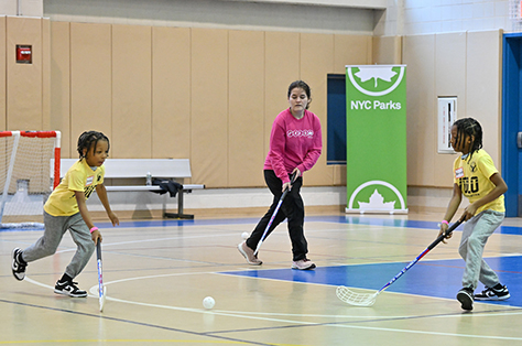 Two children wearing matching yellow shirts play street hockey on a wooden gym floor while an instructor wearing a pink NYC Parks branded shirt gives pointers.