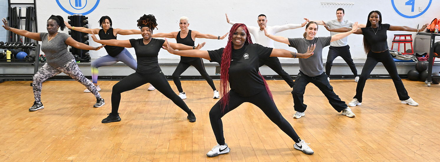 A group of individuals of different ages, genders, ethnicities, and body types stretch in the same pose together while facing the camera.