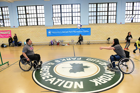 Two people in wheelchairs practice their volleyball skills in an indoor basketball court.