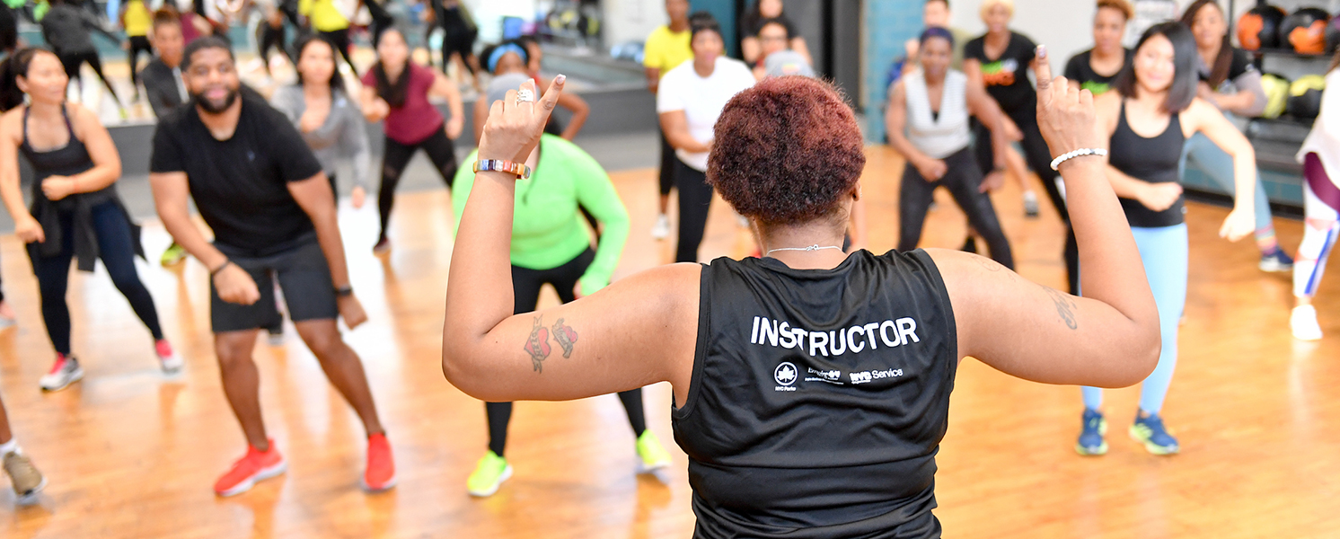 A fitness instructor holds a pose with her back to the camera, her index fingers are raised, and a class of students are following her lead. She is wearing a black shirt that reads “Instructor.”