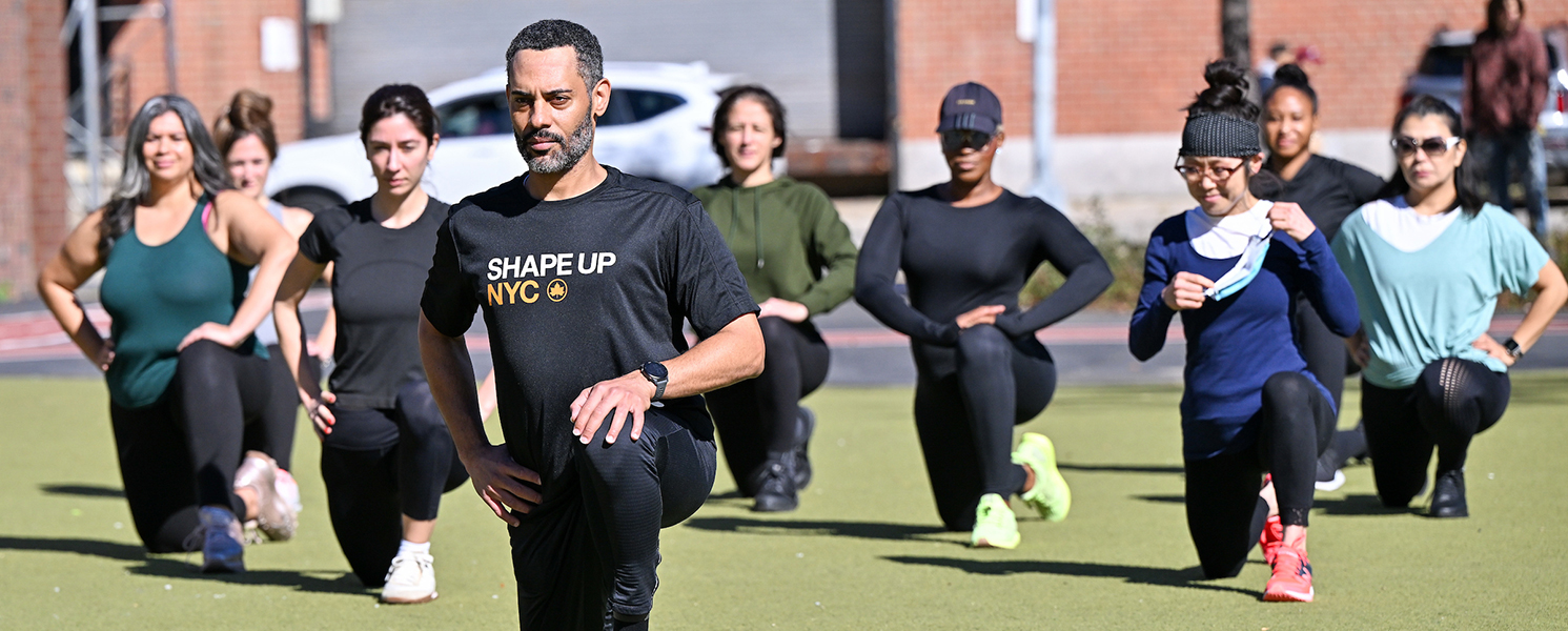 A man is kneeling with one leg outstretched while holding a determined gaze. Behind him, a class of fitness students is mirroring the fitness pose. His shirt reads “Shape Up NYC.”