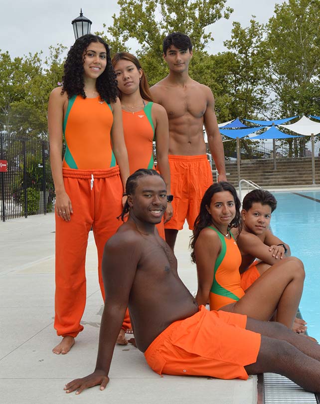 A group of six young lifeguards pose together in their orange uniforms at a city pool. They look like a close and diverse group of friends on the same team.