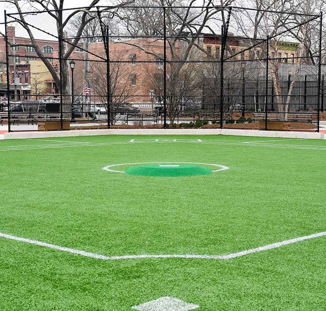 A green turf baseball diamond with a black, chainlink fence backstop with trees and buildings in the background