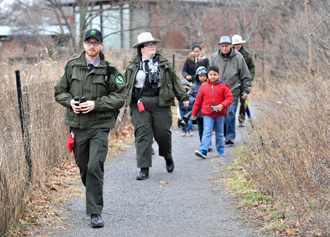 A pair of Urban Park Rangers lead a group of kids on a winter hike through tall brown marsh grasses.